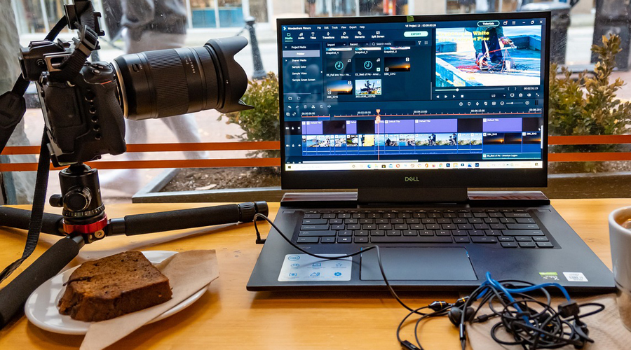 Nella foto ci sono un computer con aperto un programma di video editing. Sulla destra c'è una fotocamera e un pattino con sopra una fetta di torta. Il tutto è poggiato su un tavolo color legno.