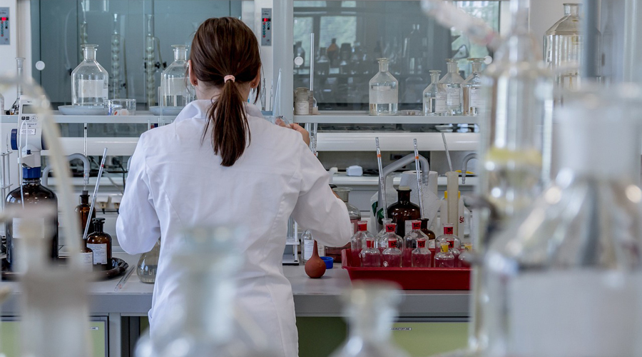 Persona di spalle con i capelli lunghi e il camice bianco in un laboratorio scientifico