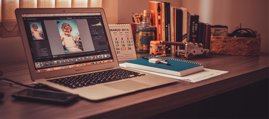 Immagine con toni caldi con un computer che ha sullo schermo aperto photoshop, sulla destra accanto al computer si trova un quaderno con penna, matita e gomma sopra. In secondo piano si trovano dei libri e un calendario.