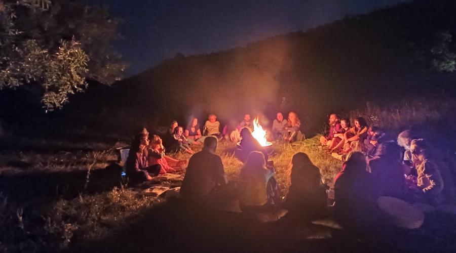 Foto notturna. Un gruppo di persone sedute in cerchio in un prato attorno a un fuoco. Sullo sfondo una collina.
