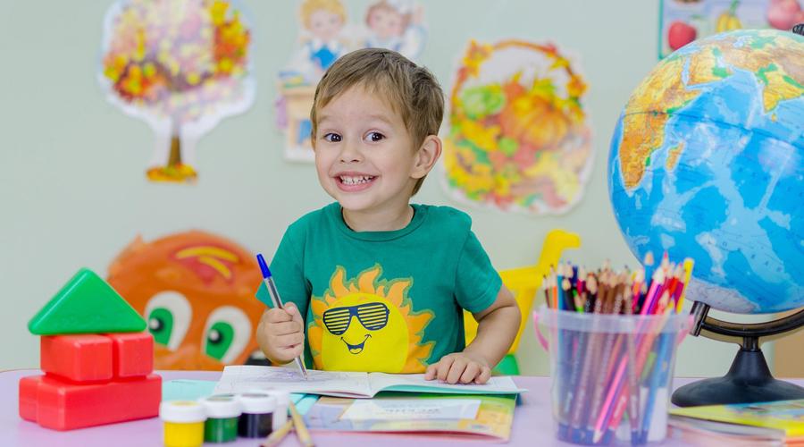 Foto di un bambino che scrive su un quaderno seduto a un tavolo. Sul tavolo ci sono colori, matite e un mappamondo.