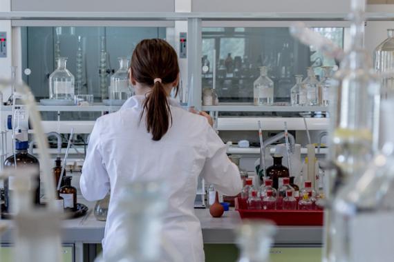 Persona di spalle con i capelli lunghi e il camice bianco in un laboratorio scientifico