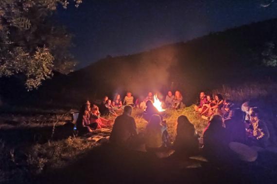 Foto notturna. Un gruppo di persone sedute in cerchio in un prato attorno a un fuoco. Sullo sfondo una collina.