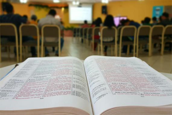 libro poggiato su un banco in primo piano, in secondo piano un gruppo di studenti che segue la lezione