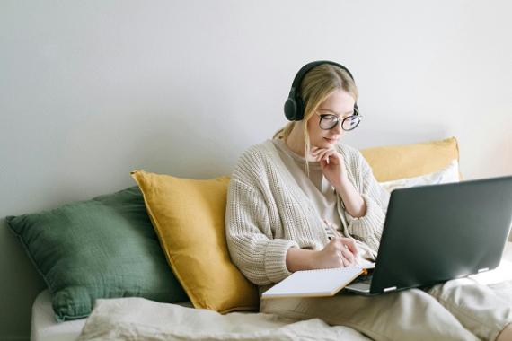 donna seduta sul letto con il pc sulle gambe e un quaderno a lato che sembra scrivere